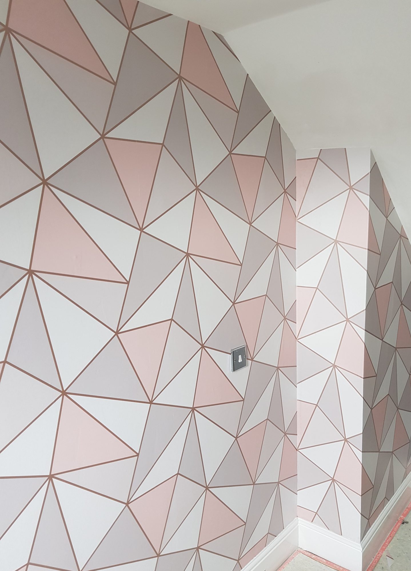 An attic bedroom papered in a pink, grey and white geometric print round the slopes and a boxed-in corner