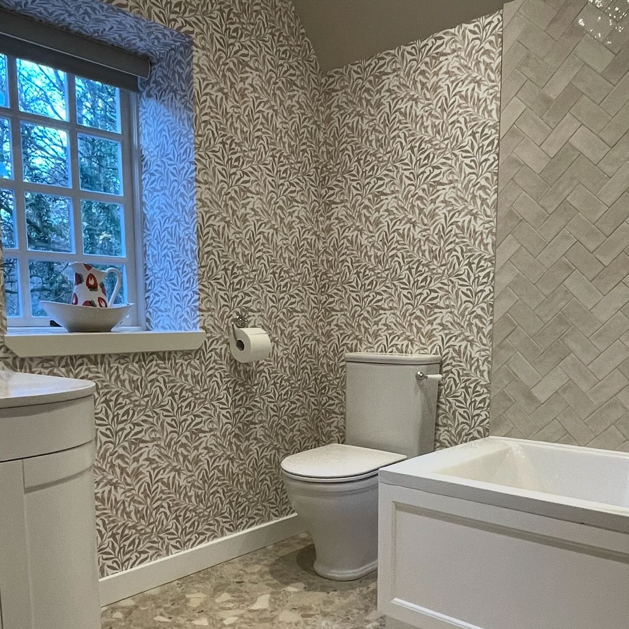 A bathroom papered in a small trailing-leaf print, into the window reveal